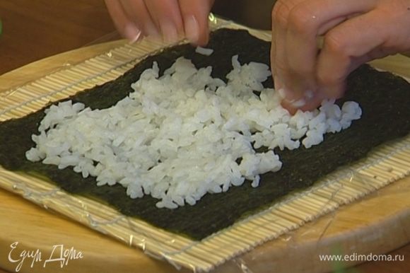 Застелить коврик для суши пищевой пленкой, положить сверху лист нори и выложить на него слой суши-риса толщиной примерно 9 мм.