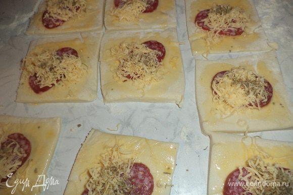 Колбасу нарезать тоненькими кружочками. Голландский сыр натереть на терке со средними отверстиями. На каждый квадратик теста выложить по кружочку колбасы, присыпать натертым сыром. Сверху слегка присыпать орегано.