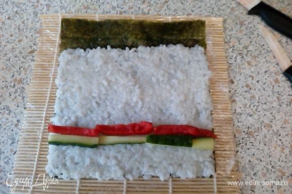 На шершавую поверхность нори выложить рис, оставив ~1–2 см листа не покрытым. Выложить бруски перца и огурца.