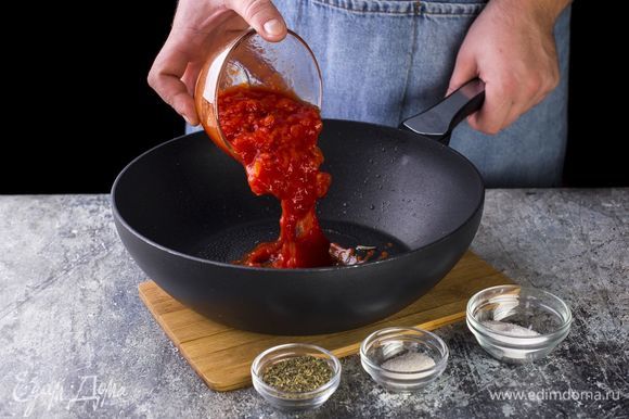 Добавьте томаты в собственном соку, соль, сахар и сухие специи. Варите на слабом огне до загустения. Чеснок и веточку базилика вытащите через 2 минуты после начала тушения.