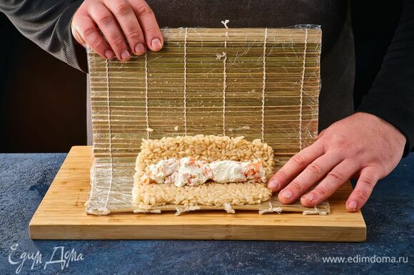 Ровным слоем выложите рис на бамбуковый коврик, обернутый в пленку. В центр рисовой массы положите начинку из рыбы и сыра. Поднимите края коврика и скатайте большой ролл.